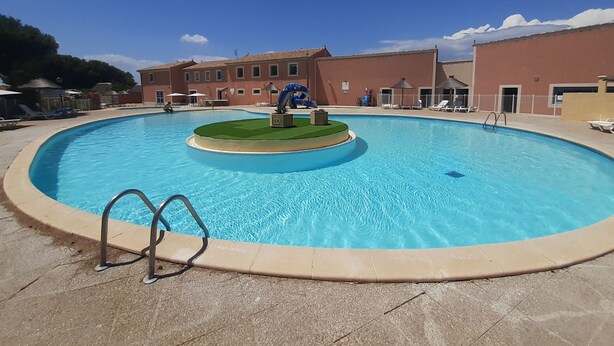 Une grande piscine extérieure dotée d’une fontaine centrale, entourée de chaises longues et bordée d’un bâtiment aux nombreuses fenêtres.