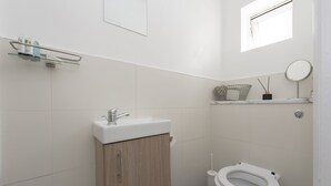Un baño pequeño y moderno con un lavamanos blanco, un mueble de madera y un inodoro. Hay una ventana que proporciona luz natural y un pequeño estante con artículos de tocador.