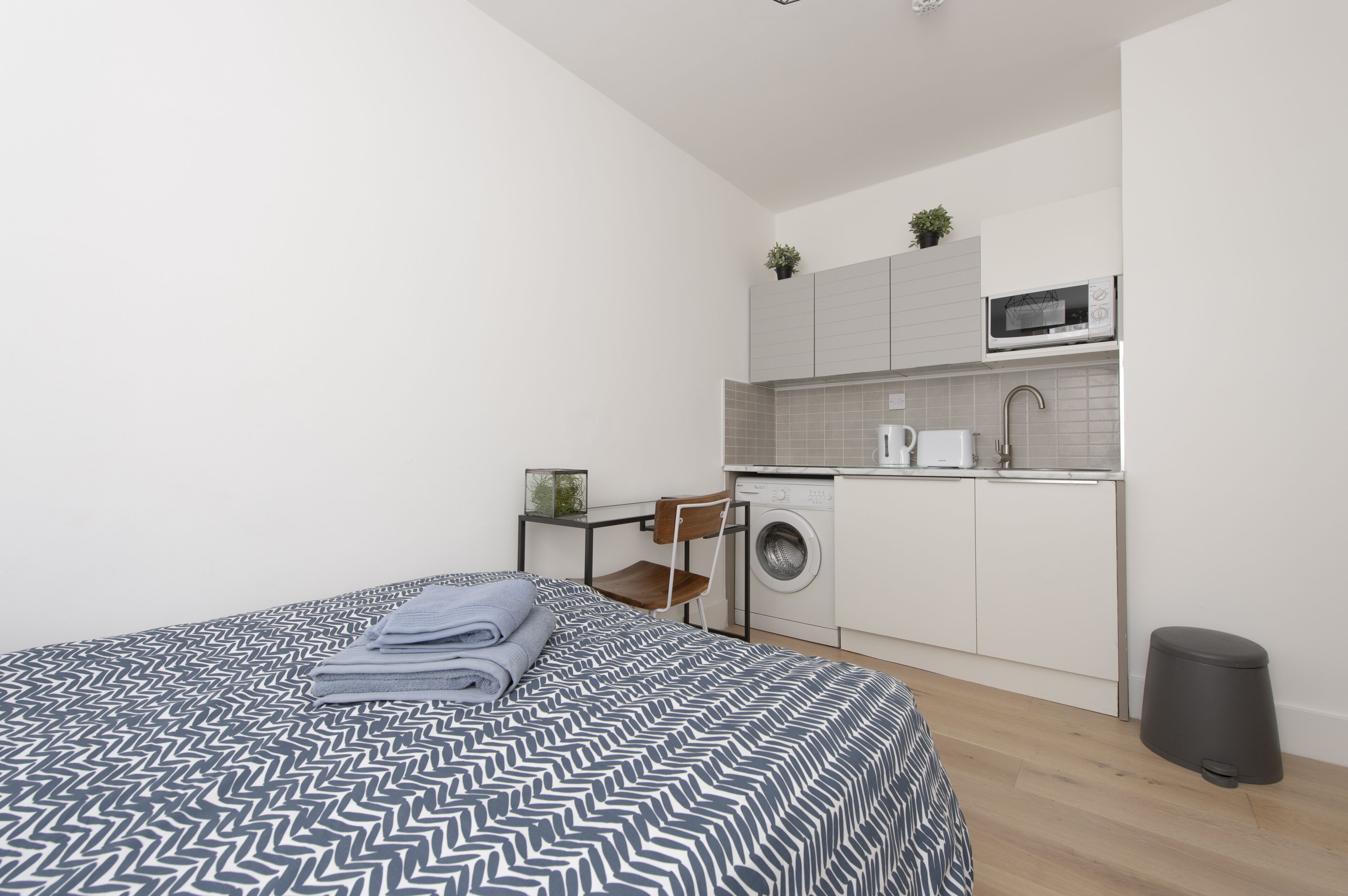 A modern, compact room with a neatly made bed, a washing machine, and a small kitchenette.