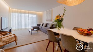 A modern living room with a sofa, a dining table with chairs, a coffee table, and a large window with sheer curtains.