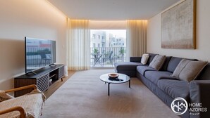 A modern living room with a sofa, a coffee table, and a TV. There is a balcony with a view of buildings.