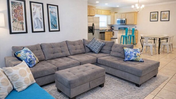 A living room with a sectional sofa, ottoman, and a blue couch. There is a kitchen in the background with a dining area. - Oversized 2 Bedroom - Modern - Pool Near Beach (South Padre Island)