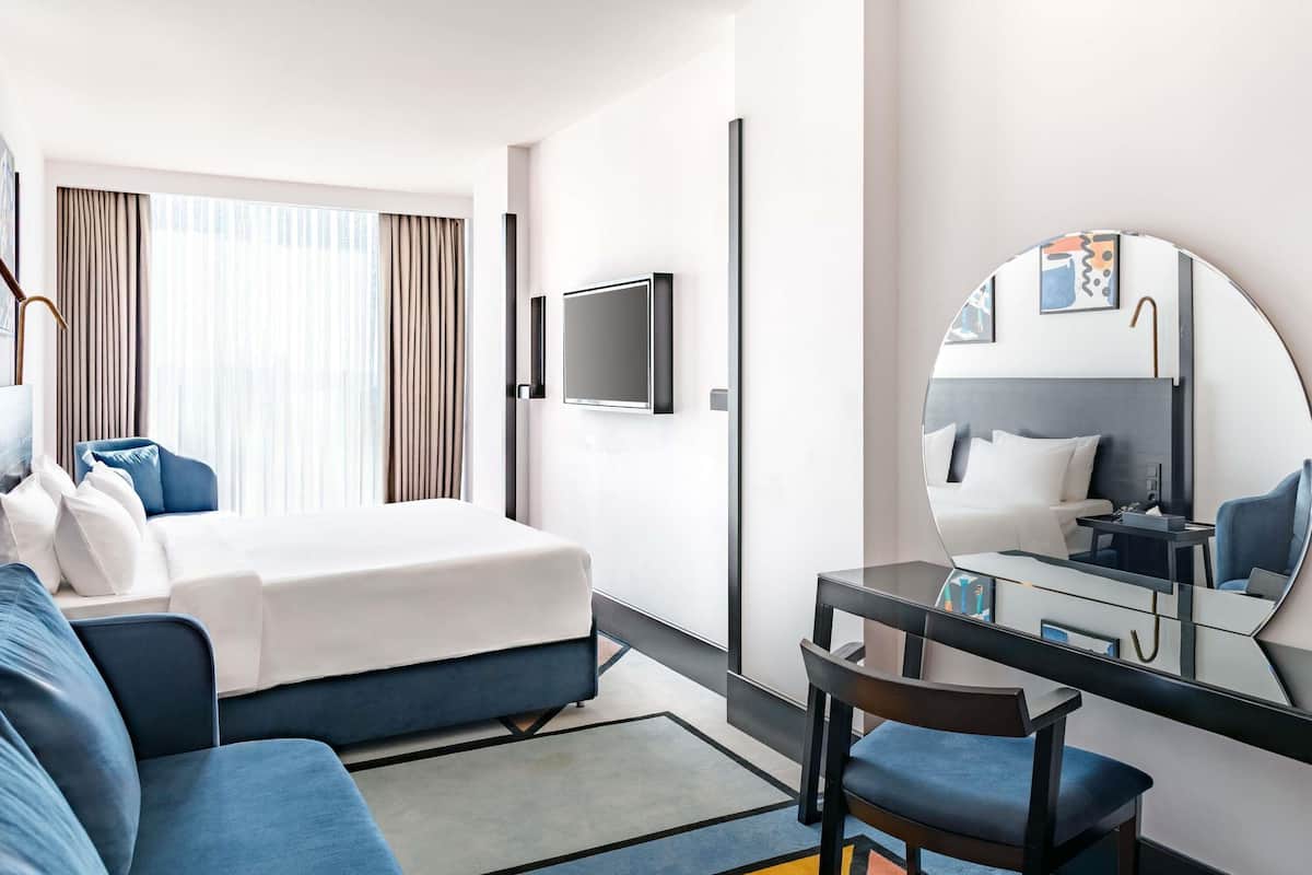A hotel room with a bed, a desk with a chair, a television, a large mirror, and a window with curtains.