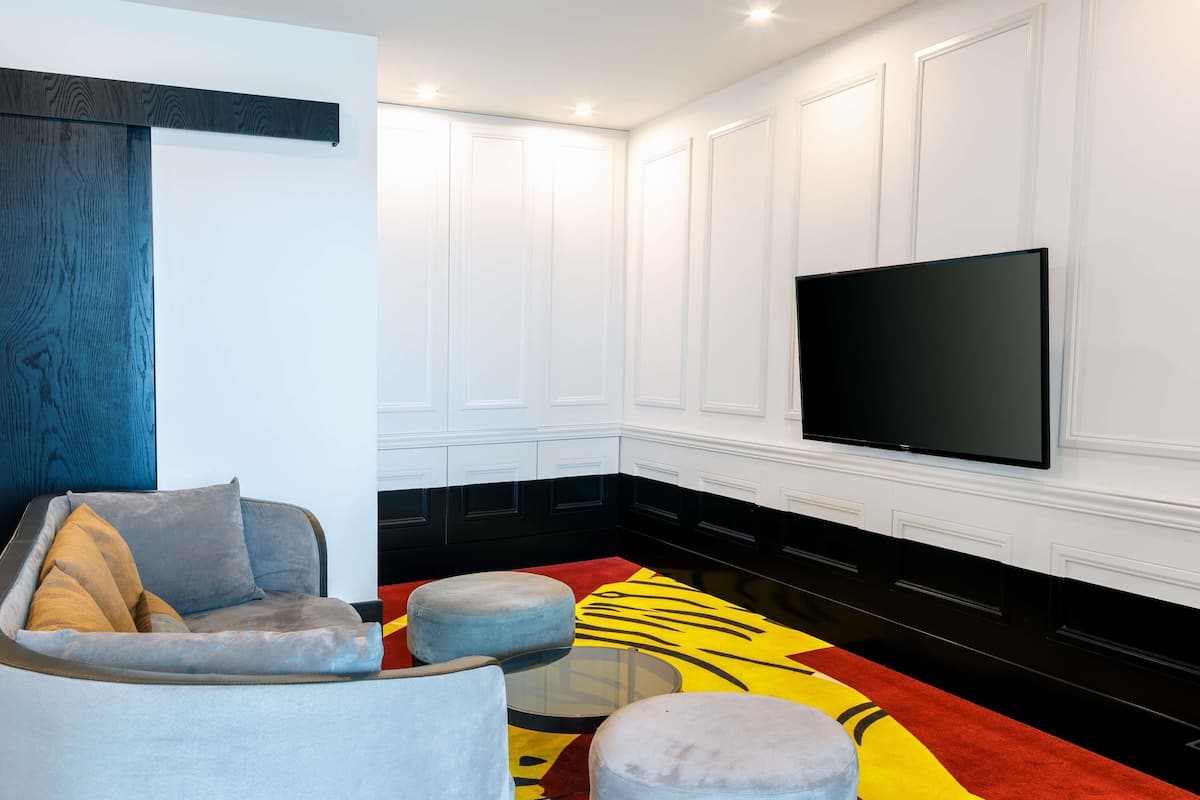 A modern living room with a flat-screen TV, a grey sofa, ottomans, and a patterned rug.