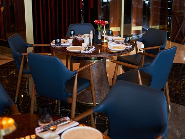 A round dining table with blue chairs, a vase with red flowers, and plates set with cutlery.