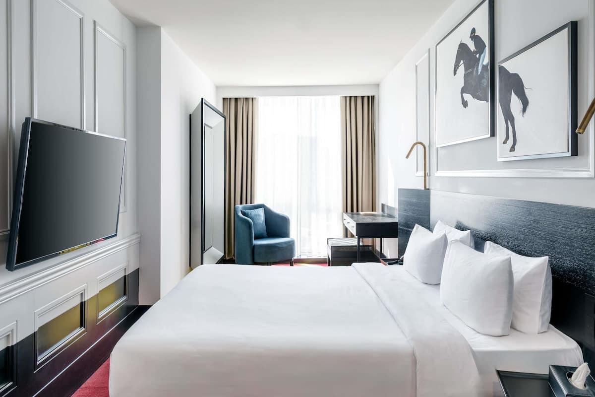 A hotel room with a large bed, a TV, a desk with a lamp, a chair, and a window with curtains.