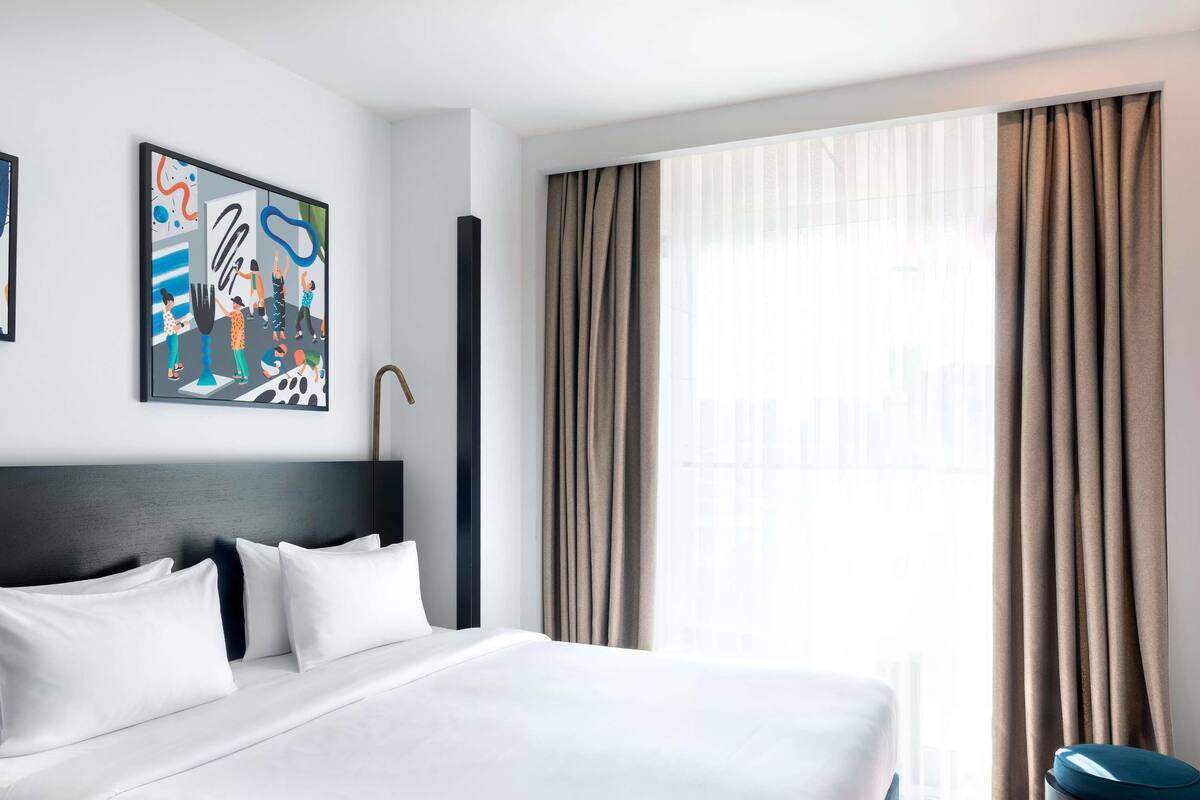 A hotel room with a large bed, a headboard, a window with curtains, and a painting on the wall.