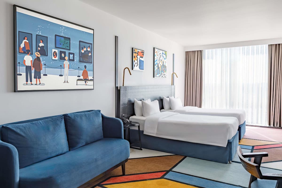 A hotel room with a blue sofa, a bed with white bedding, a colorful rug, and artwork on the walls.