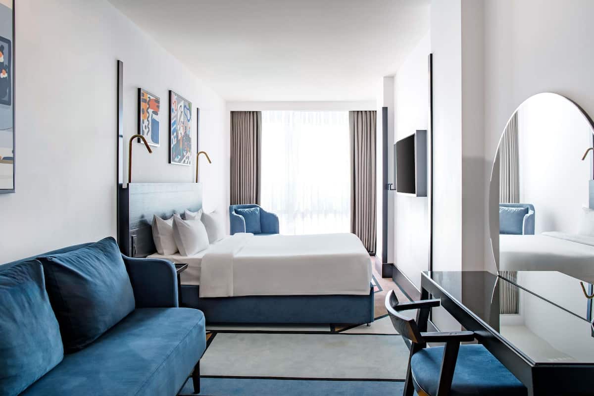 A hotel room with a bed, a sofa, a desk with a chair, a TV, and a large mirror.