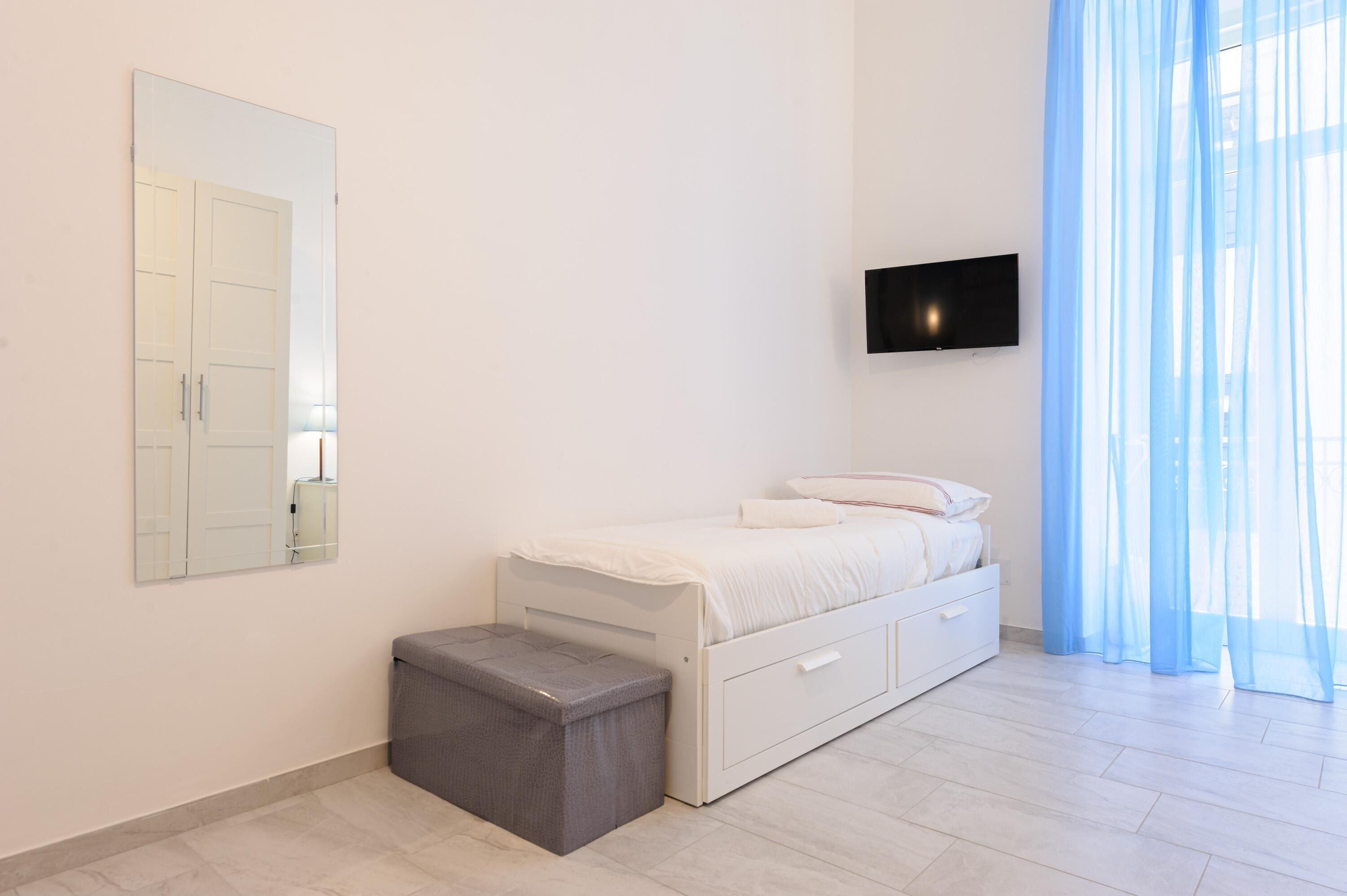 Une chambre à coucher moderne avec un lit, un petit banc, une télévision et des rideaux légers.