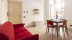A modern living room with a red sofa, a dining table with four chairs, and a small bench. There are two wall-mounted shelves with plants, a door, and a window with a view of buildings. - Palazzo Nan 3 Piano Scala C (Naples)