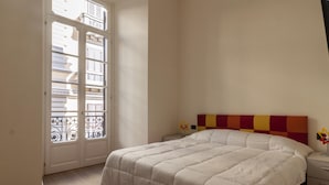 A bedroom with a large bed, a nightstand, a window with a view of buildings, and a door with a metal gate. - Palazzo Nan 3 Piano Scala C (Naples)