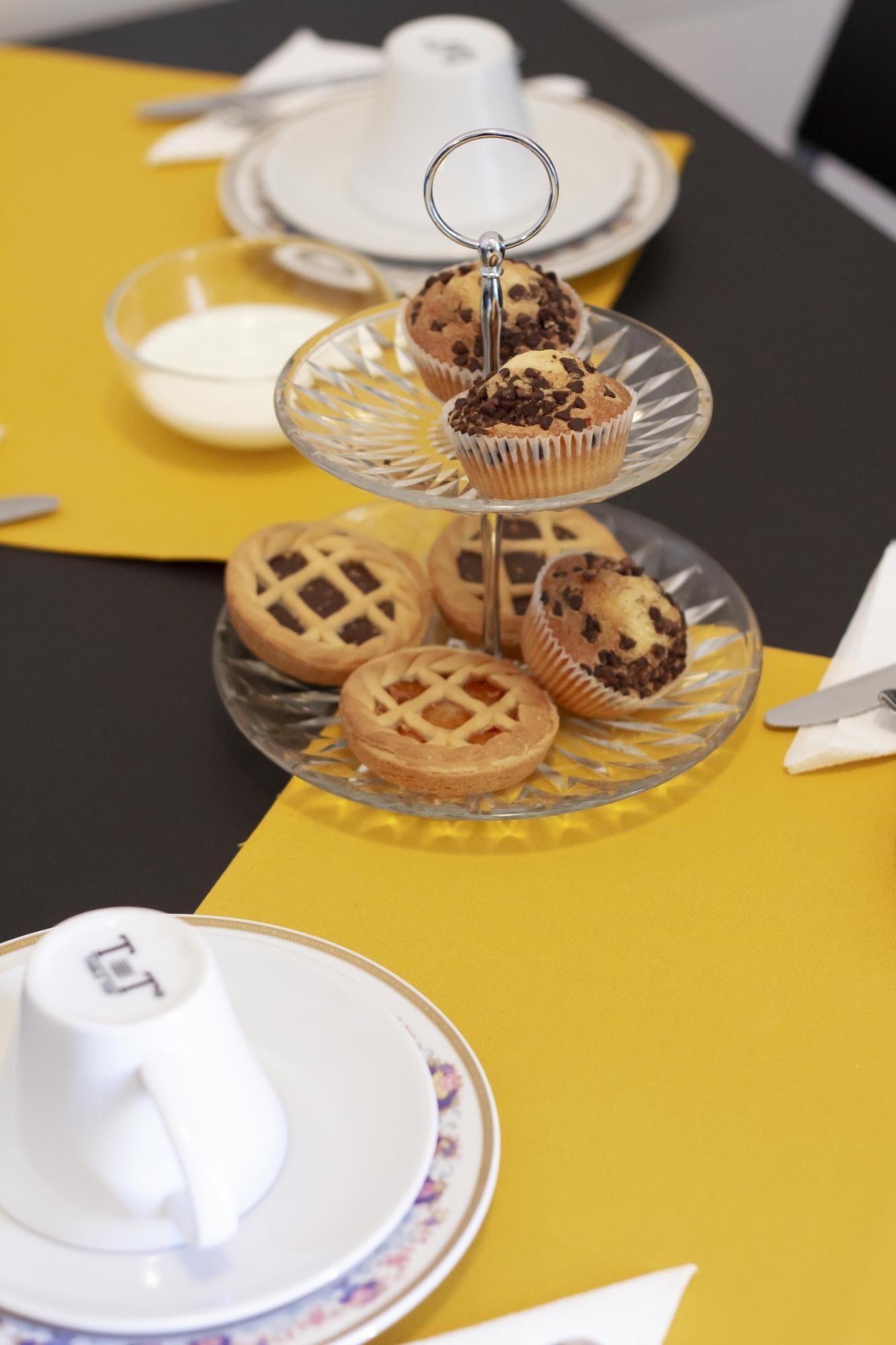 Une table recouverte d’une nappe jaune, avec une théière blanche et un présentoir en verre à plusieurs niveaux, sur lequel sont disposés des biscuits et un muffin.