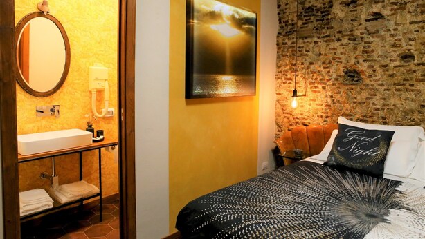 A bathroom with a sink, mirror, and towel rack. There is a bed with a black and white patterned bedspread and a pillow with the text "Good Night". The room has a brick wall and a hanging light fixture. - Napolicentrico in Napoli (Naples)