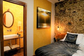 A bathroom with a sink, mirror, and towel rack. There is a bed with a black and white patterned bedspread and a pillow with the text "Good Night". The room has a brick wall and a hanging light fixture. - Napolicentrico in Napoli (Naples)