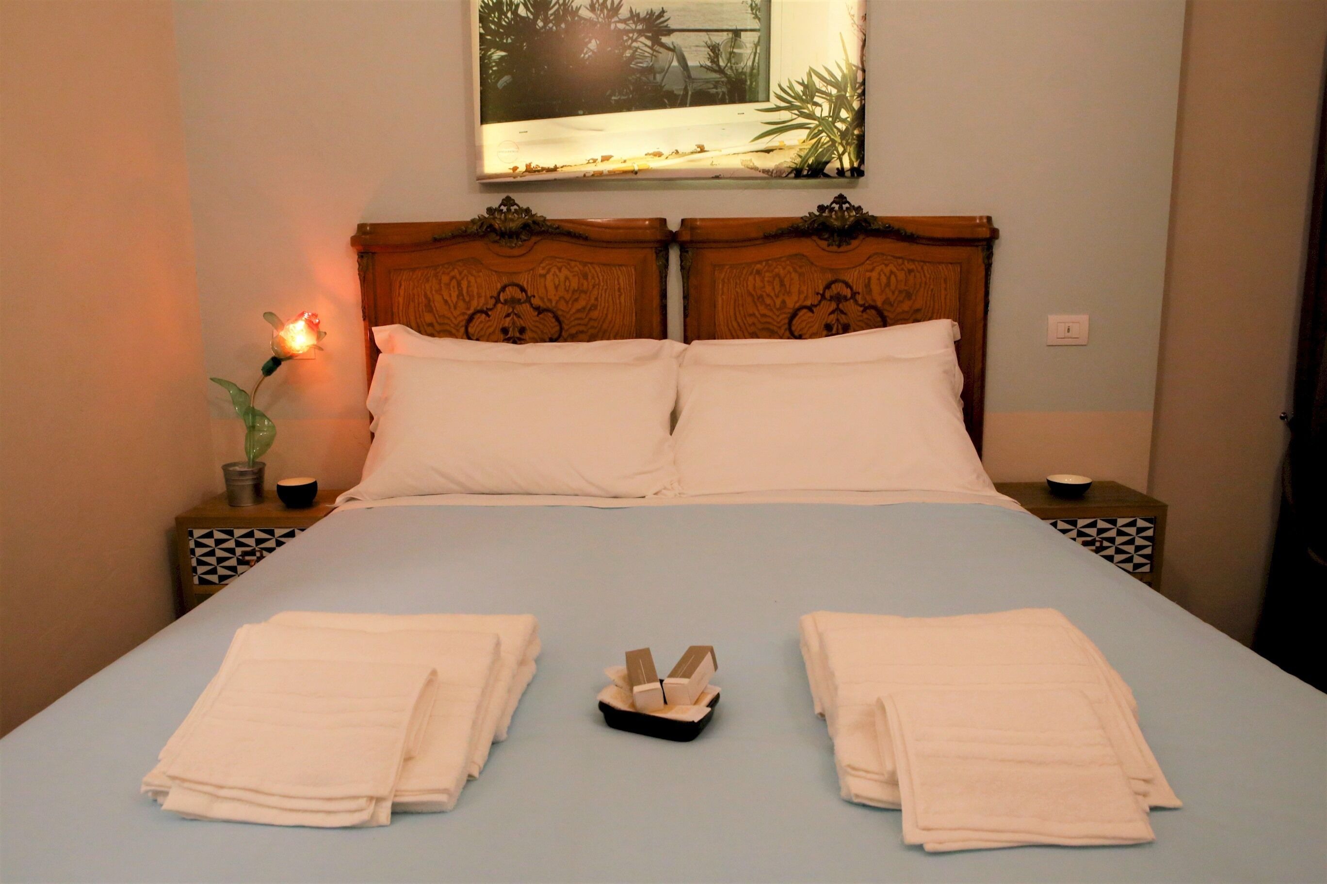 A double bed with white linens, two bedside tables with geometric-patterned nightstands, a small potted plant, and a framed picture on the wall.