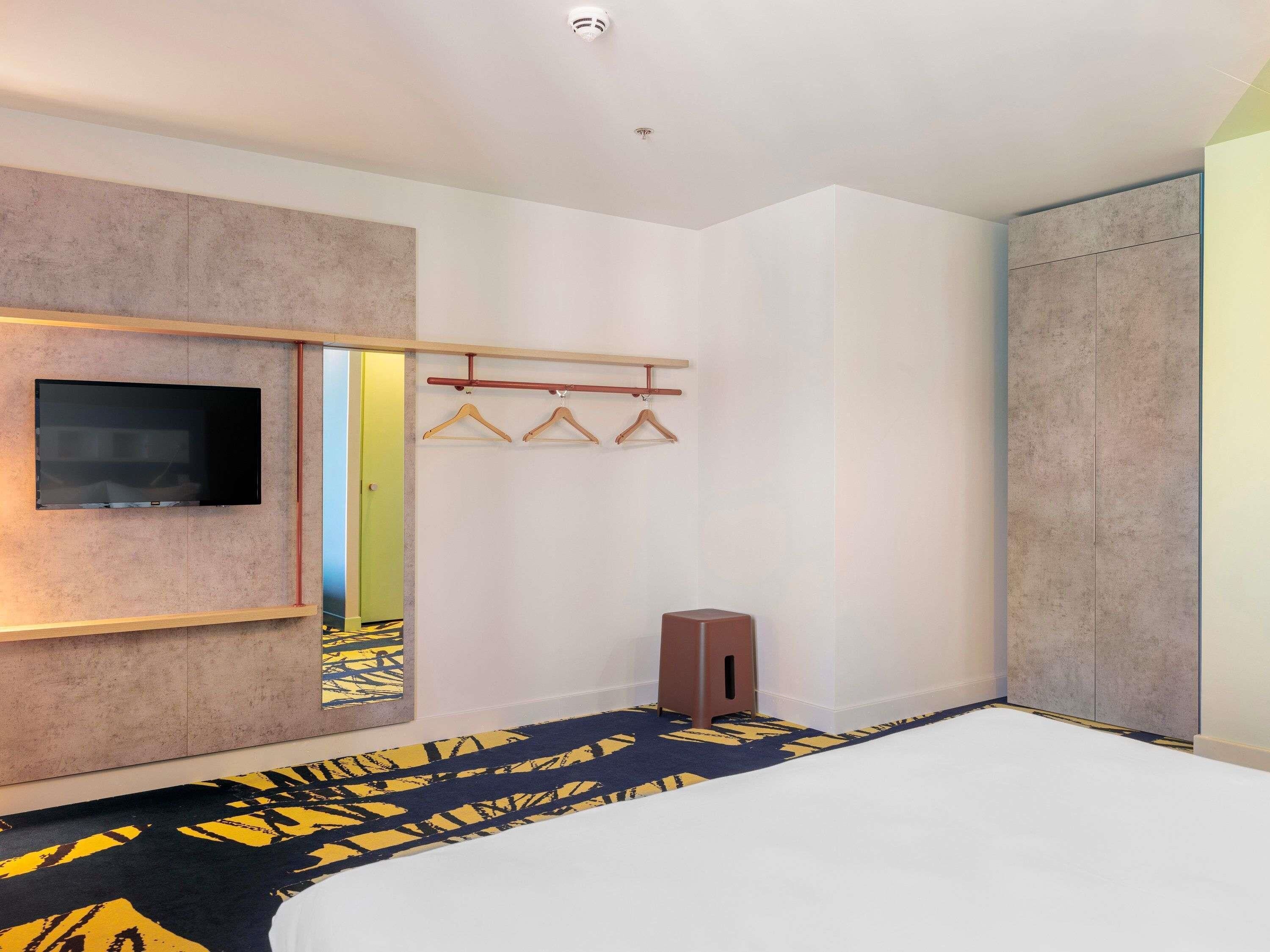 A hotel room with a bed, a TV mounted on the wall, a coat rack with hangers, a small stool, and a door leading to another room.