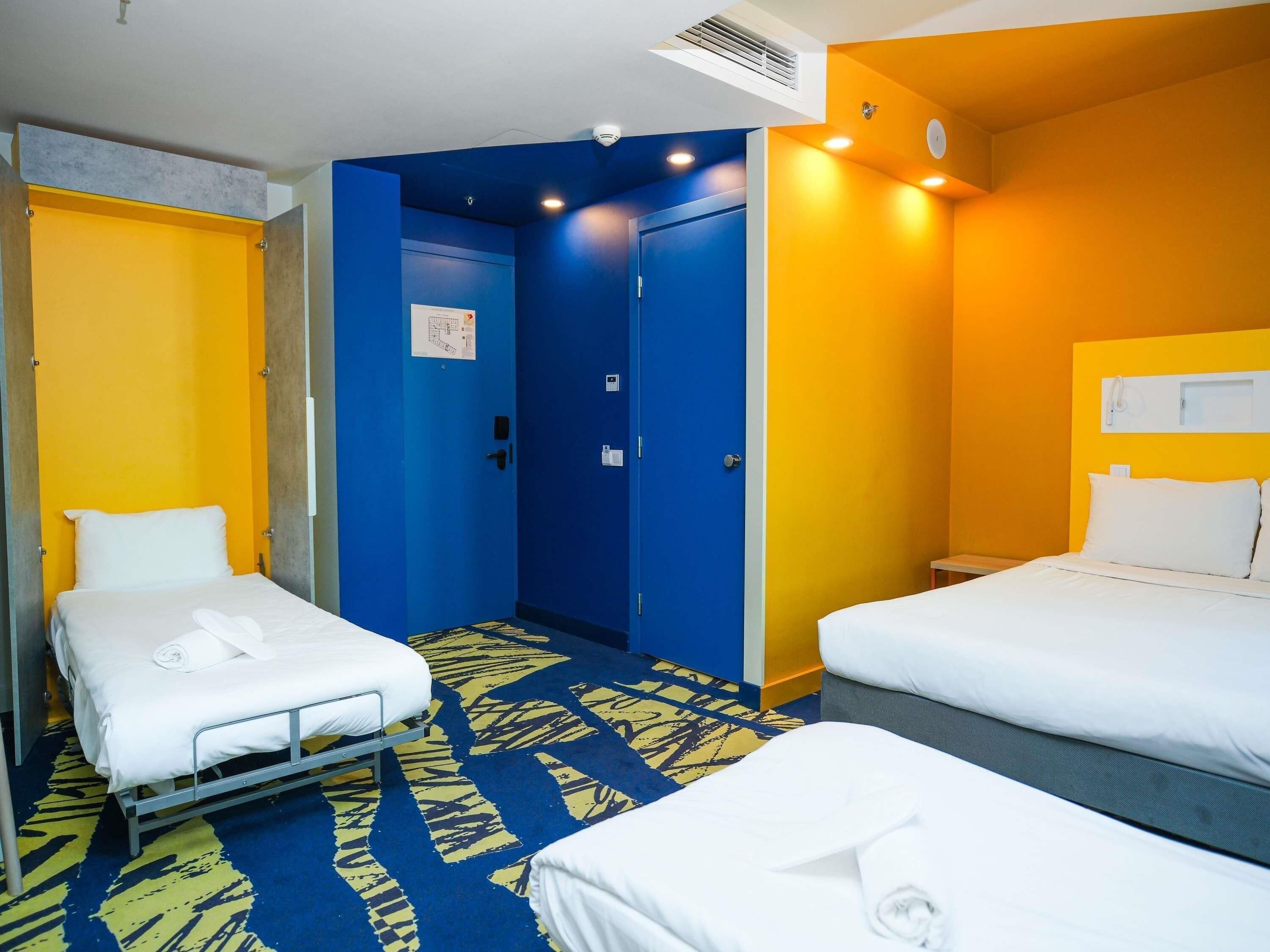 Une chambre d’hôtel avec deux lits, une porte bleue et un mur d’accent jaune.