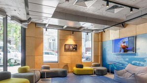 A modern lobby with seating areas, a large window with a view of the street, and a mounted artwork. - Ibis Budget Tbilisi Center (Tbilisi)