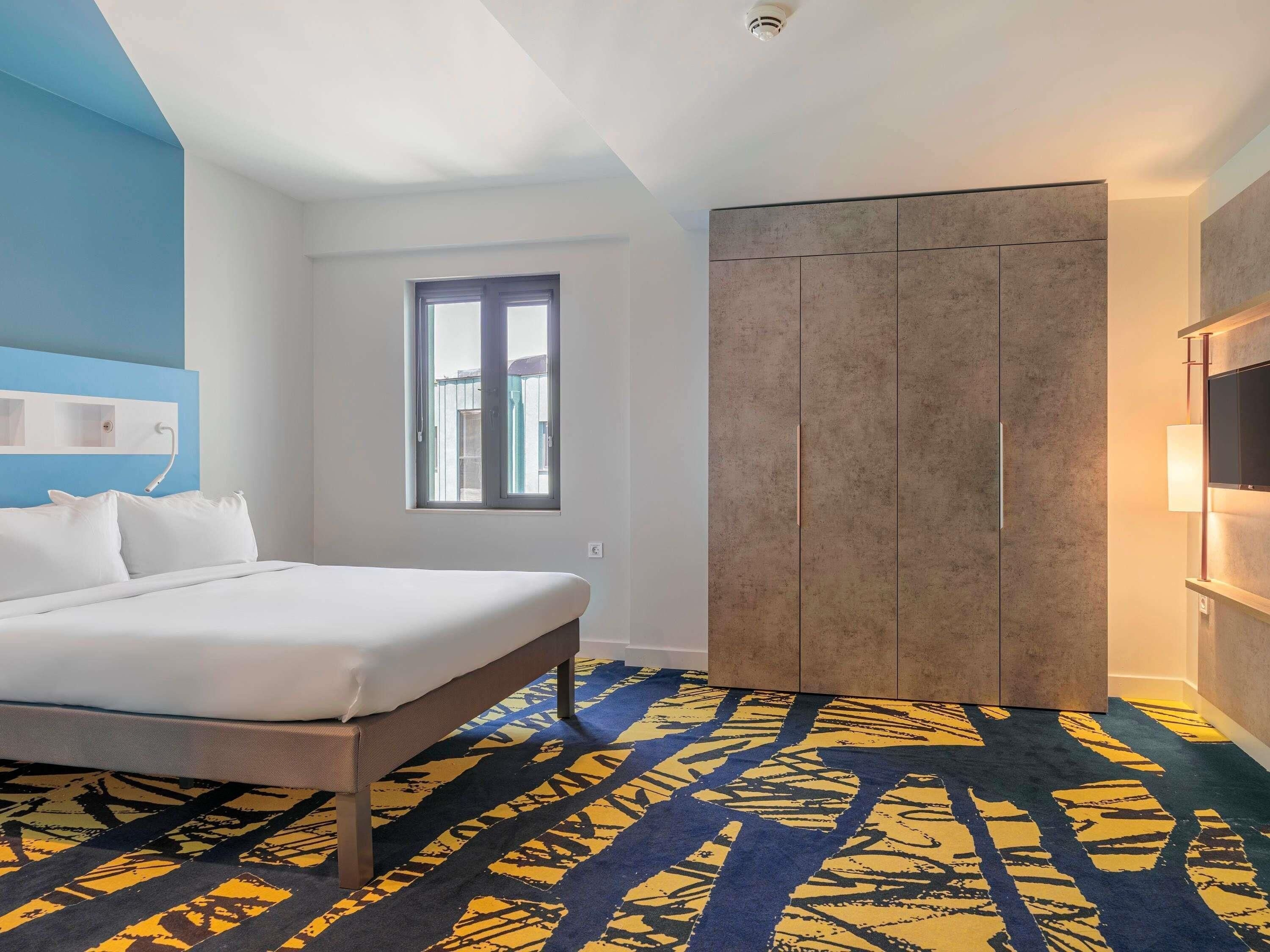 A hotel room with a bed, a wardrobe, a window, and a patterned carpet.