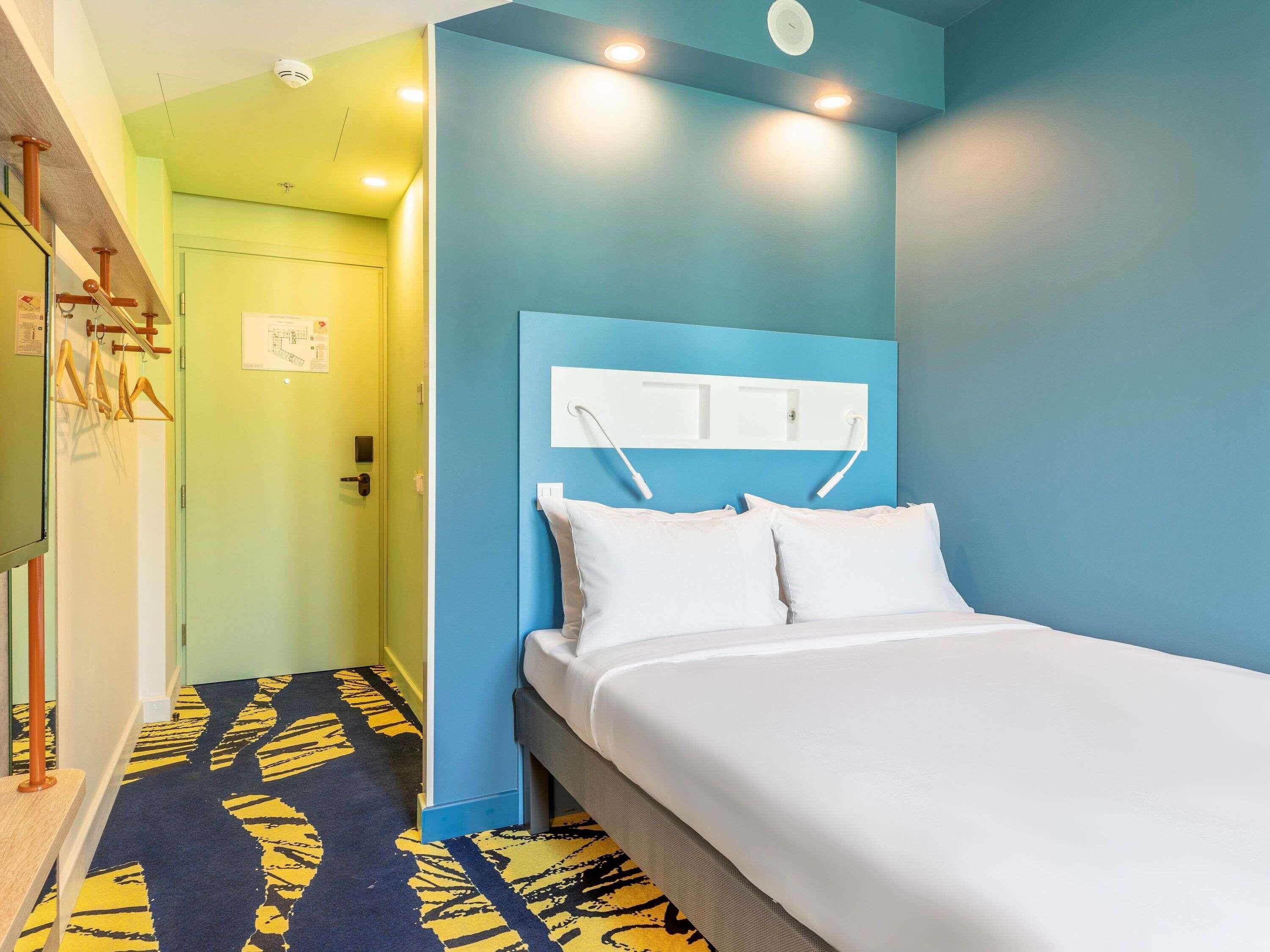 A hotel room with a bed, a blue accent wall, a patterned carpet, and a hallway with a green door.