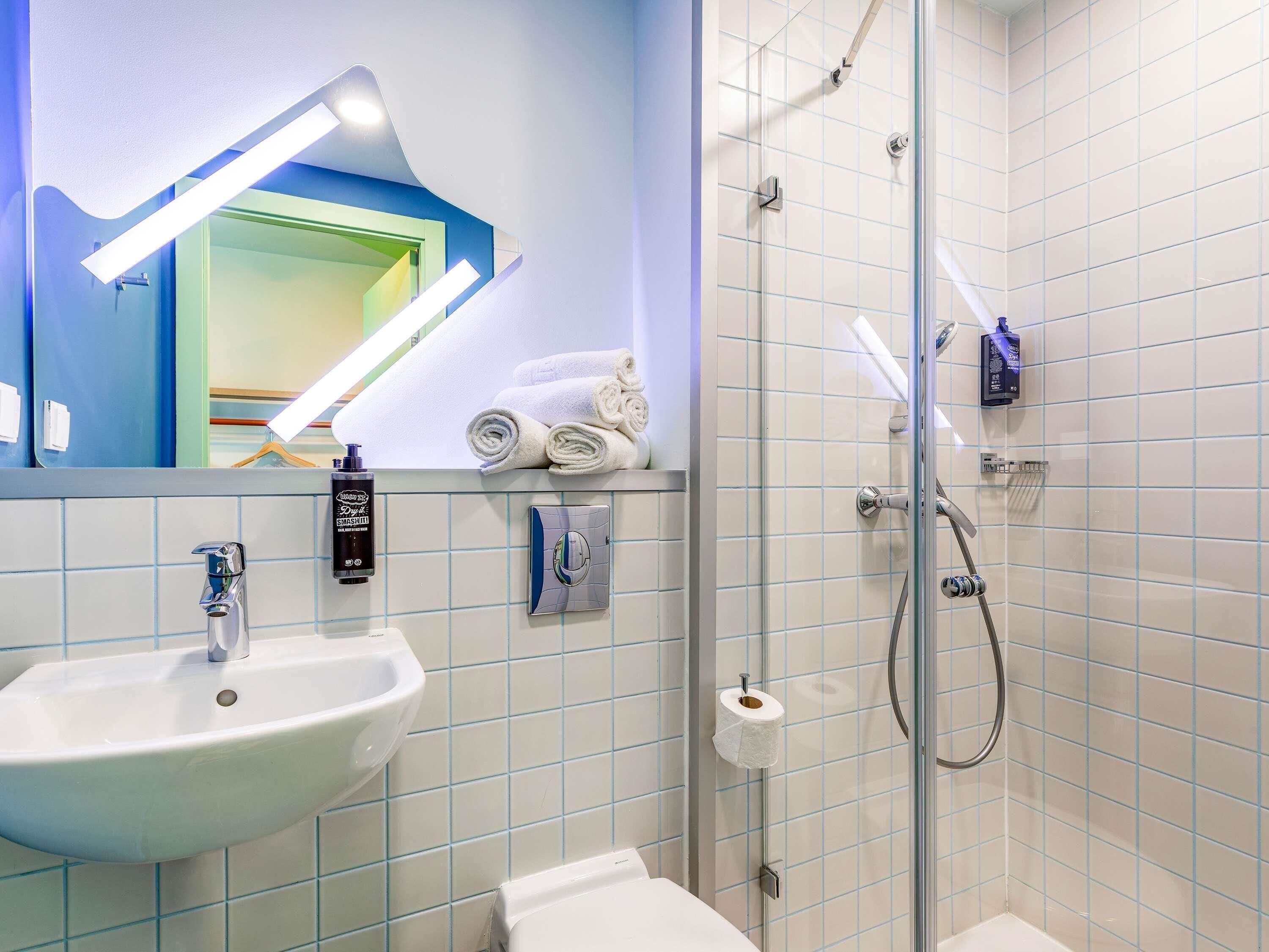A bathroom with a sink, mirror, towel rack, shower, and toilet paper.