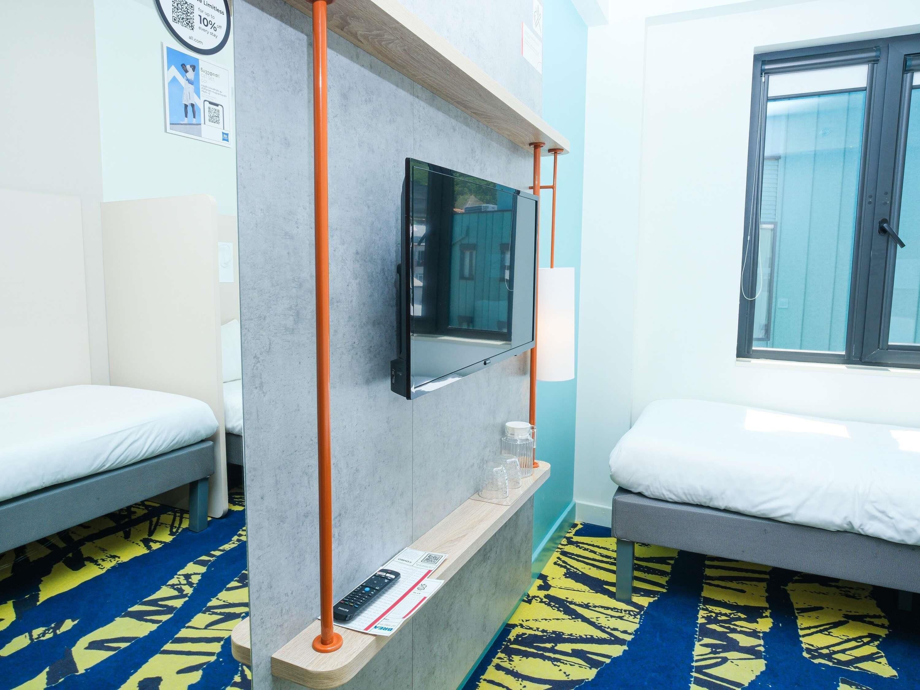 A hotel room with two single beds, a TV mounted on the wall, a window with a view, and a blue patterned carpet.