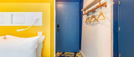 A hotel room with a bed, a wall socket, a door, and a hallway with a patterned carpet.