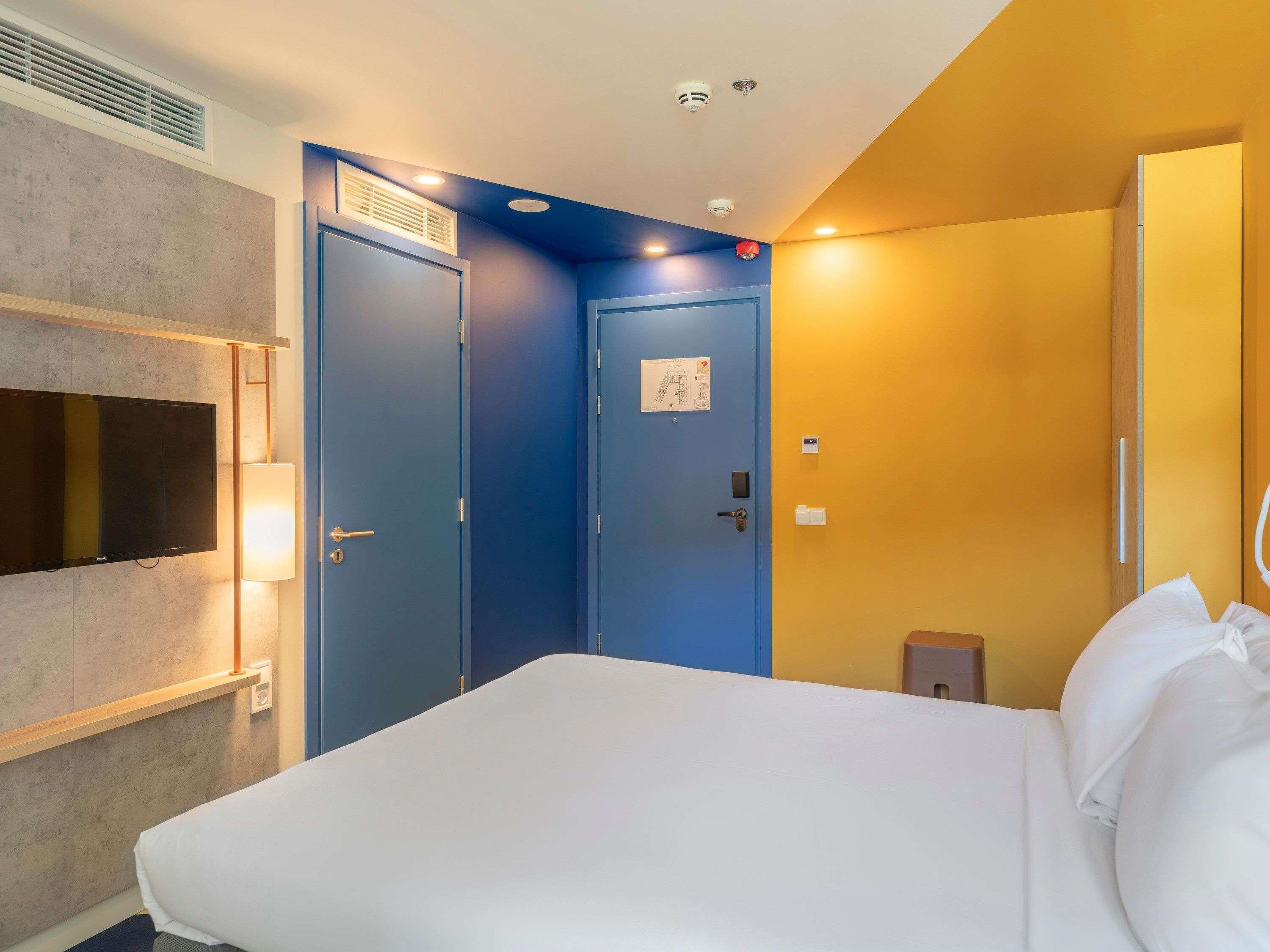 Une chambre d’hôtel avec un lit, une télévision, une lampe fixée au mur et une porte dotée d’une serrure.