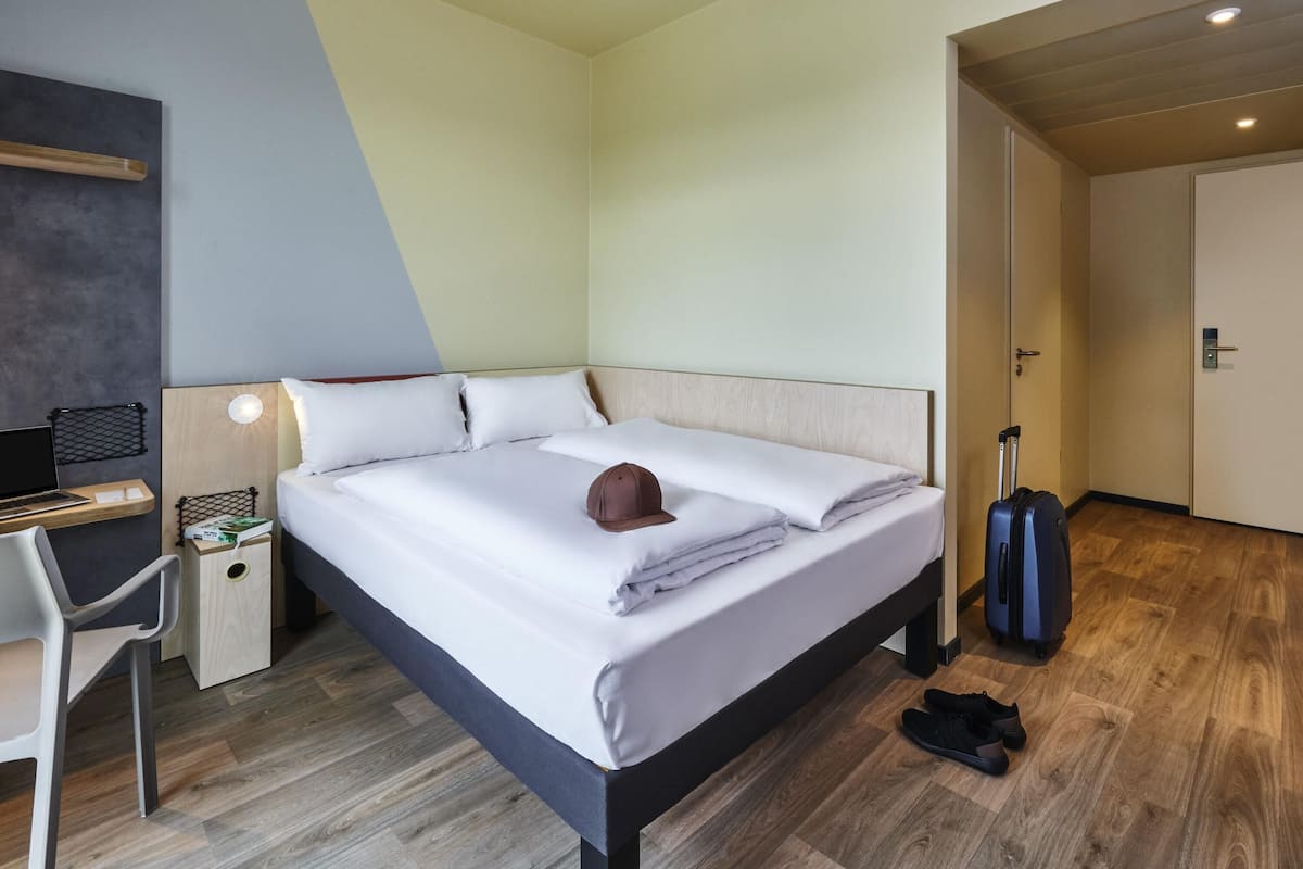 A modern hotel room with a bed, desk, chair, and a suitcase.
