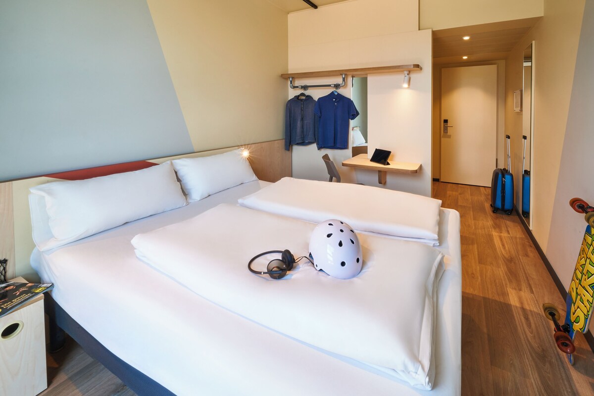 A double bed with white bedding, a white helmet, and headphones on the bed. There is a wooden nightstand with a magazine on it.