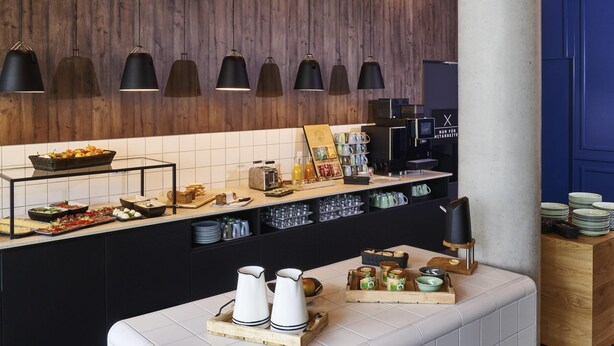 A modern kitchen with a wooden wall, black pendant lights, and a buffet counter with various food items. - Ibis Budget Singen (Singen)