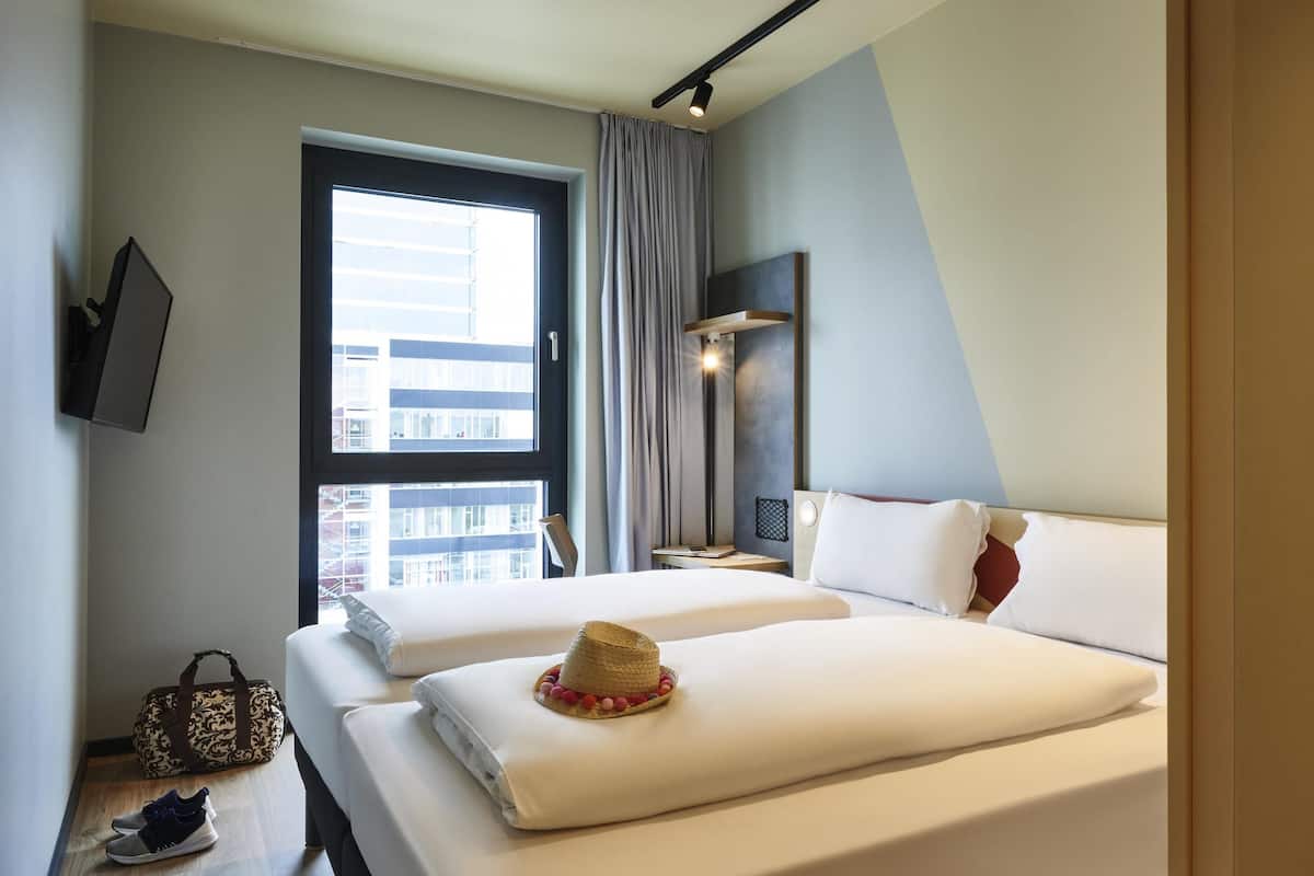 A hotel room with two beds, a large window, a bedside table with a lamp, and a view of buildings outside.