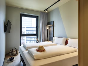 A hotel room with two beds, a large window, a bedside table with a lamp, and a view of buildings outside. - Ibis Budget Singen (Singen)
