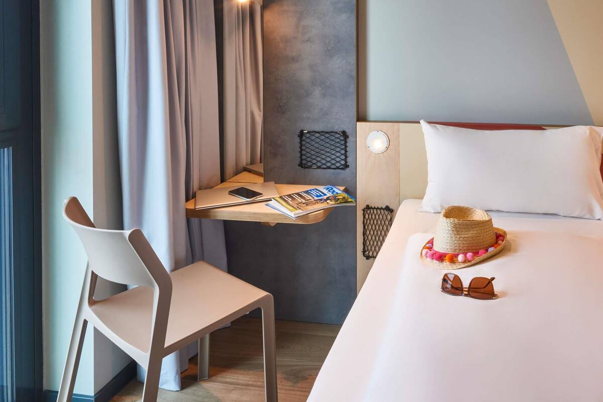 A hotel room with a bed, a desk with a laptop, a chair, and a bedside table with a hat and sunglasses.