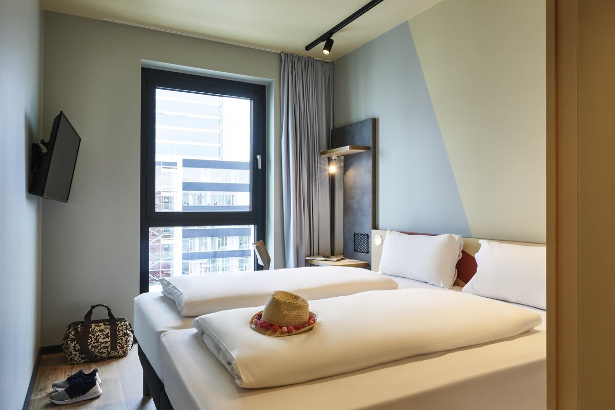 A hotel room with two beds, a large window, a bedside table with a lamp, and a view of buildings outside.