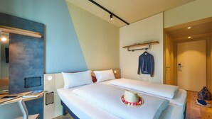 A hotel room with two beds, a desk, and a chair. There is a hat on the bed and a jacket on the wall. - Ibis Budget Singen (Singen)