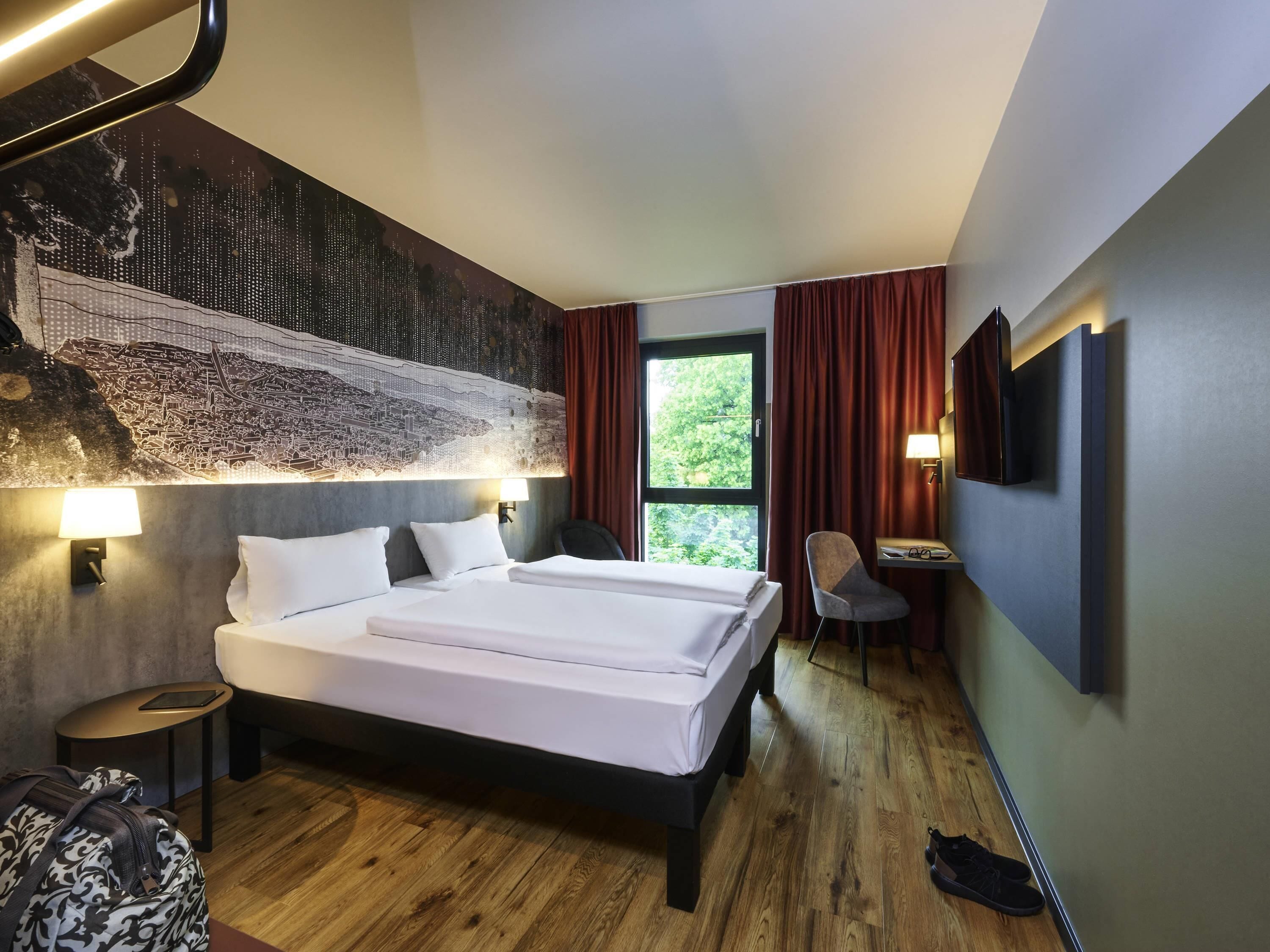 Una habitación de hotel moderna con una cama grande, un escritorio con silla, un televisor y un mural con vista a la ciudad.