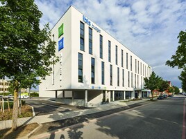 A modern building with the 'ibis Budget' sign, featuring a flat facade, large windows, and a parking area with a red line marking.