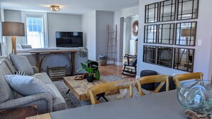 A living room with a sofa, coffee table, and chairs. There is a television, a plant, and a ladder in the background. - Luxury living, quick walk to town. King bed. (Eastport)