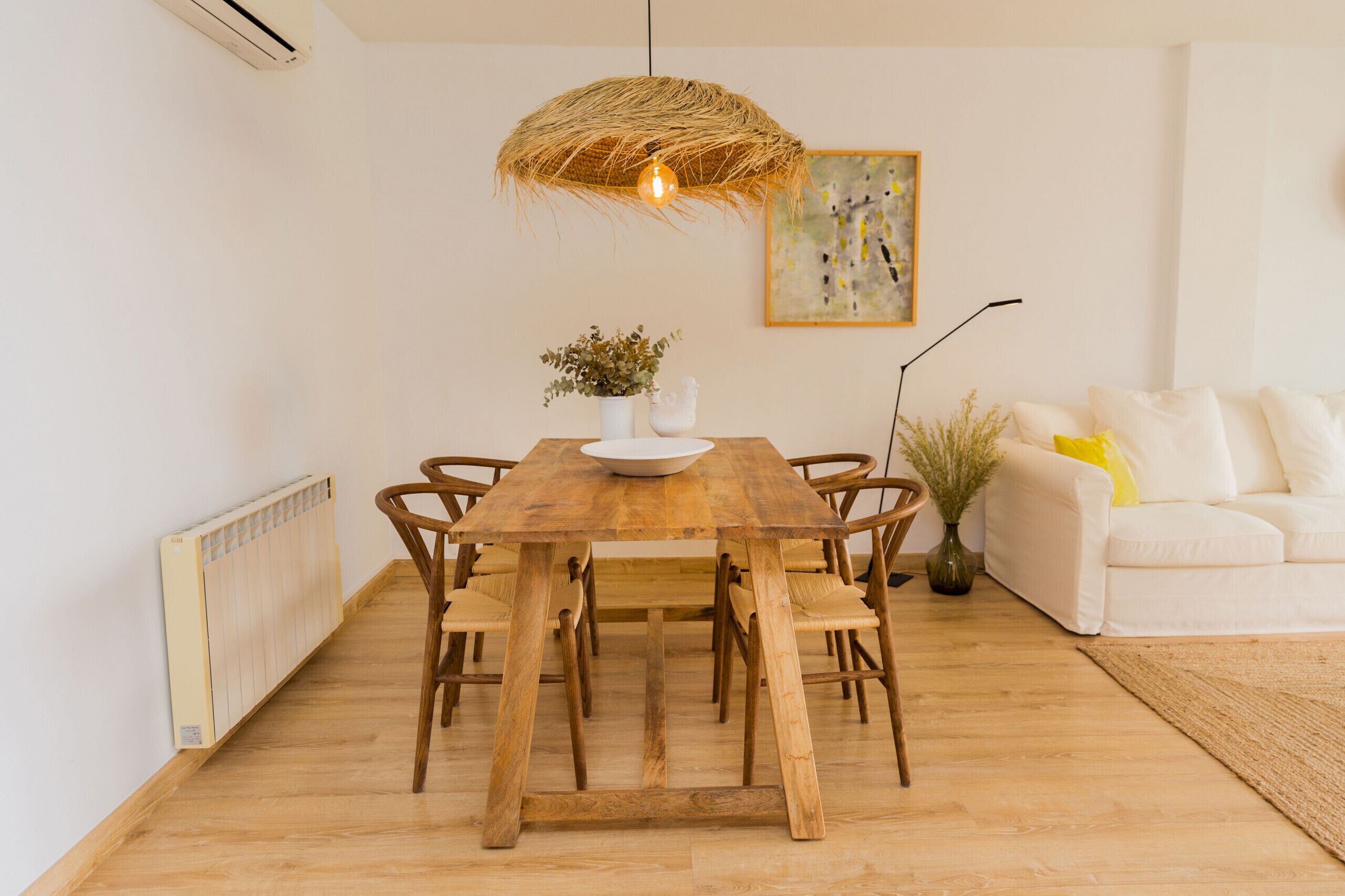 A dining area with a wooden table, chairs, a hanging light fixture, a white vase with plants, and a white sofa with a yellow pillow.