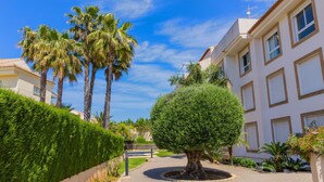 Un camino pavimentado que conduce a un edificio con setos cuidados y un pequeño árbol.
