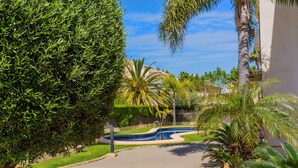 Un camino pavimentado que lleva a una piscina rodeada de palmeras y un edificio al fondo.