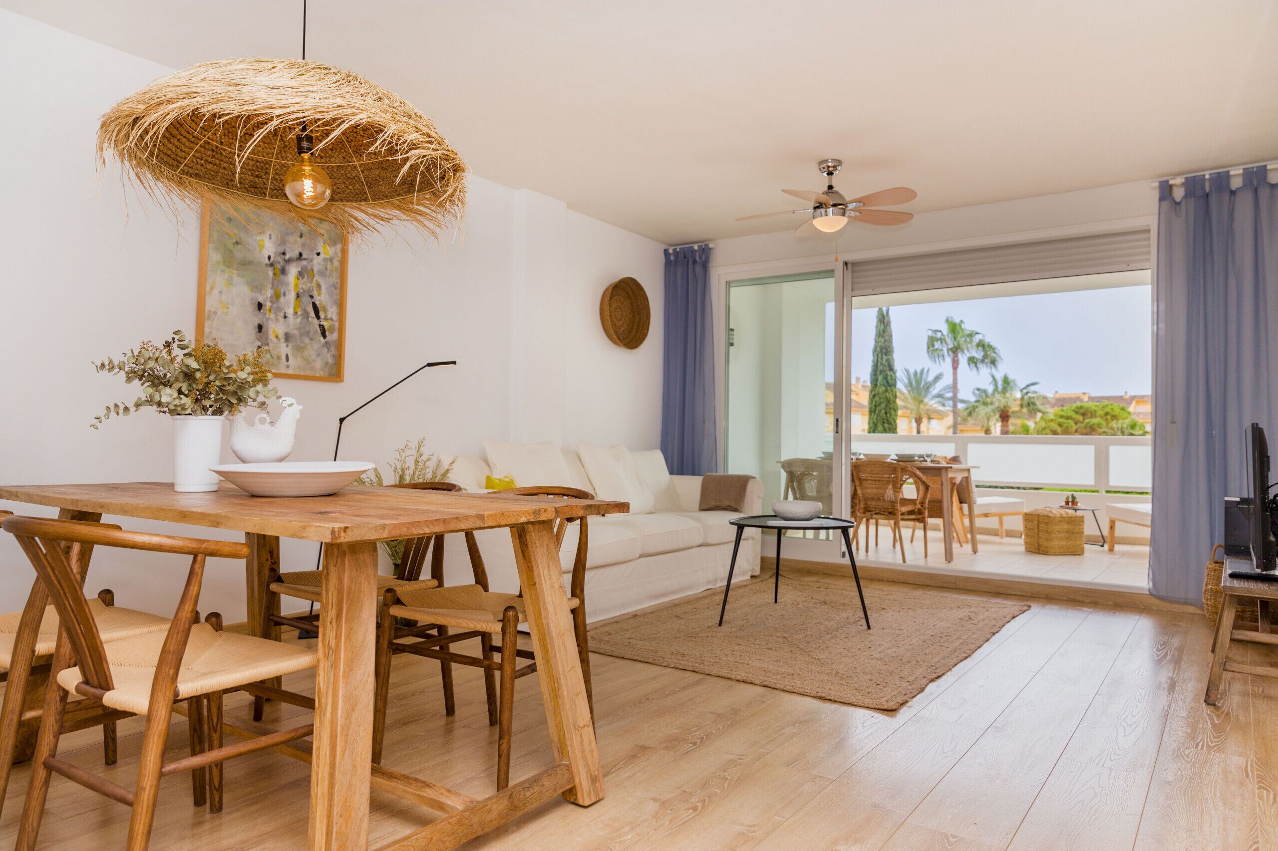 A spacious living area with a wooden dining table, a sofa, and a balcony with a view of palm trees.