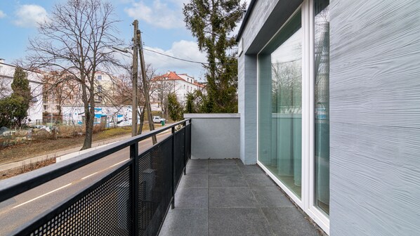 En balkon med gelænder i sort metal, flisegulv og udsigt til en gade med træer og bygninger.