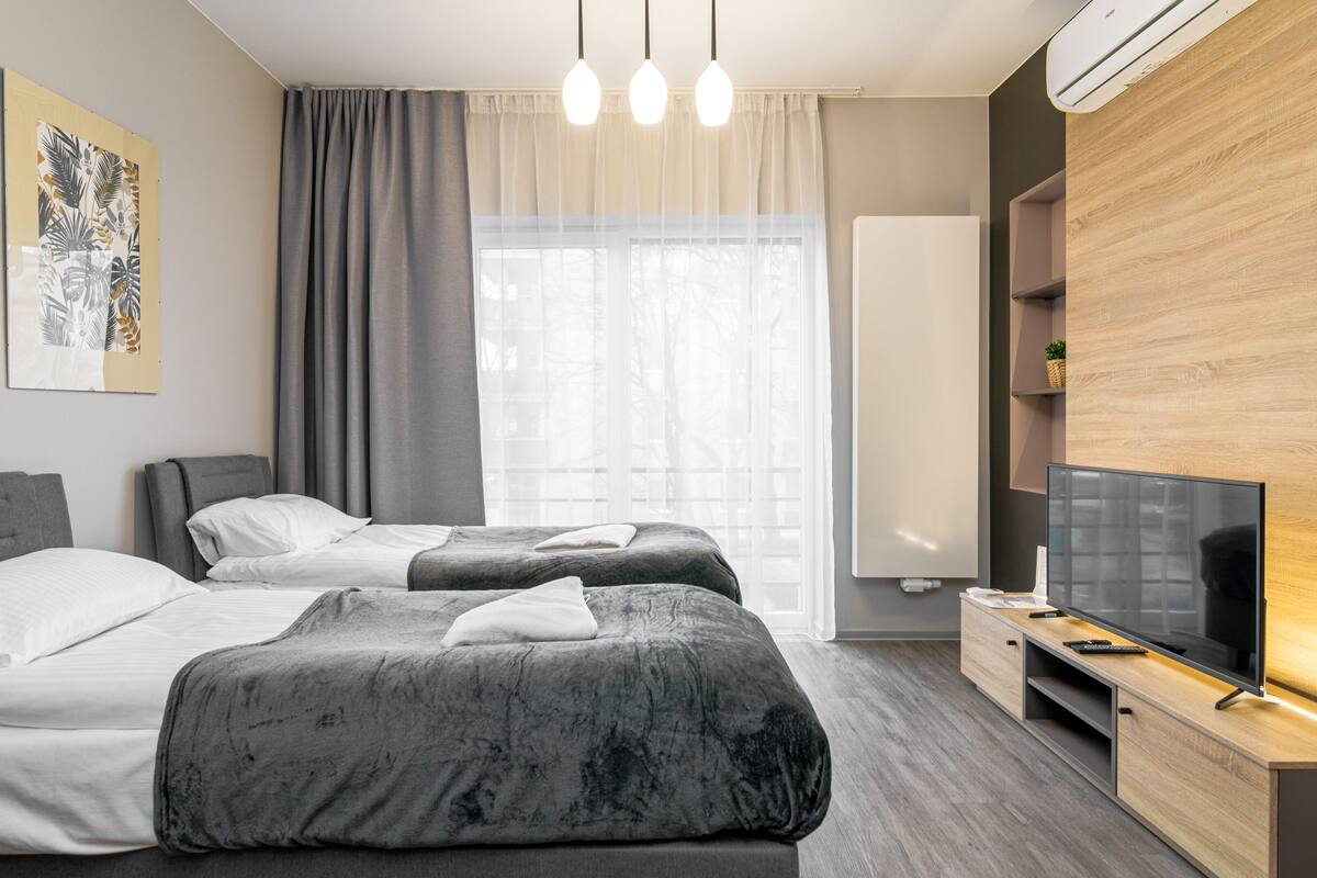 Ein modernes Hotelzimmer mit zwei Betten, einem Flachbildfernseher, einem Holzschrank und einem großen Fenster mit Vorhängen.