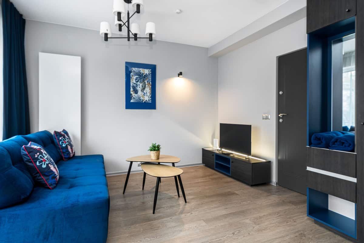 Ein modernes Wohnzimmer mit einem blauen Sofa, einem kleinen runden Tisch mit Pflanze, einem Flachbildfernseher und einem dunkelholzigen Kleiderschrank.