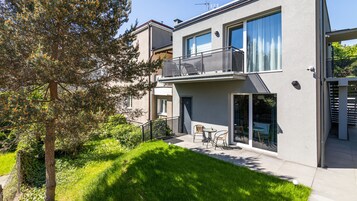 A modern two-story building with a balcony, a small patio with outdoor seating, and a well-maintained garden.