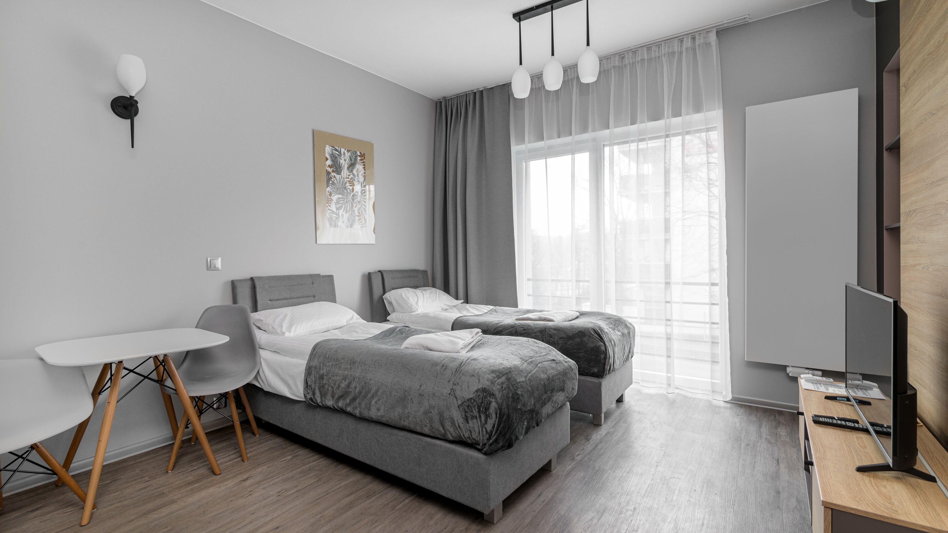 A modern hotel room with two beds, a small table, a chair, a TV, and a large window with curtains.