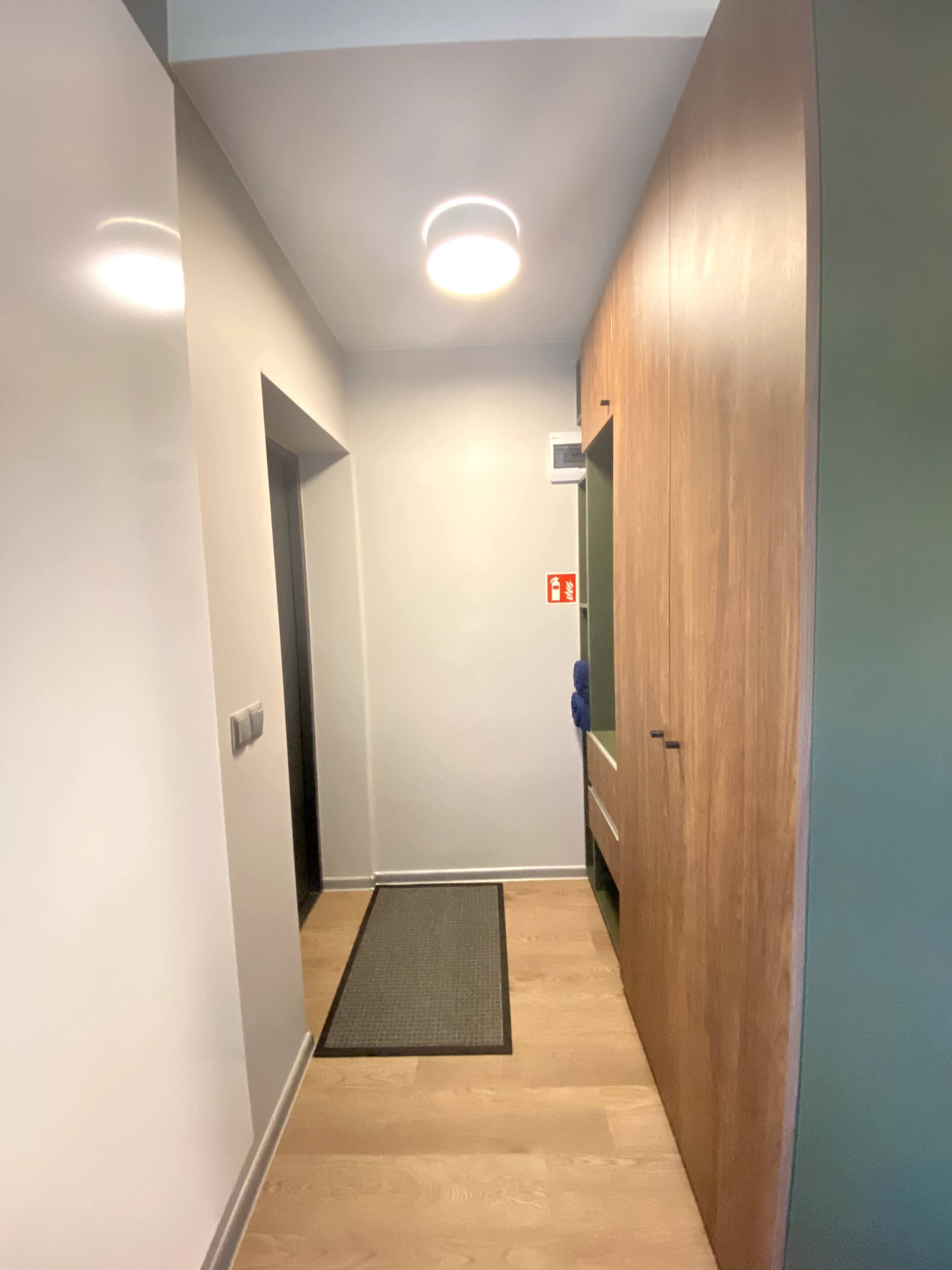 Un pasillo estrecho con una puerta de madera, un felpudo pequeño en la entrada y una lámpara en el techo.